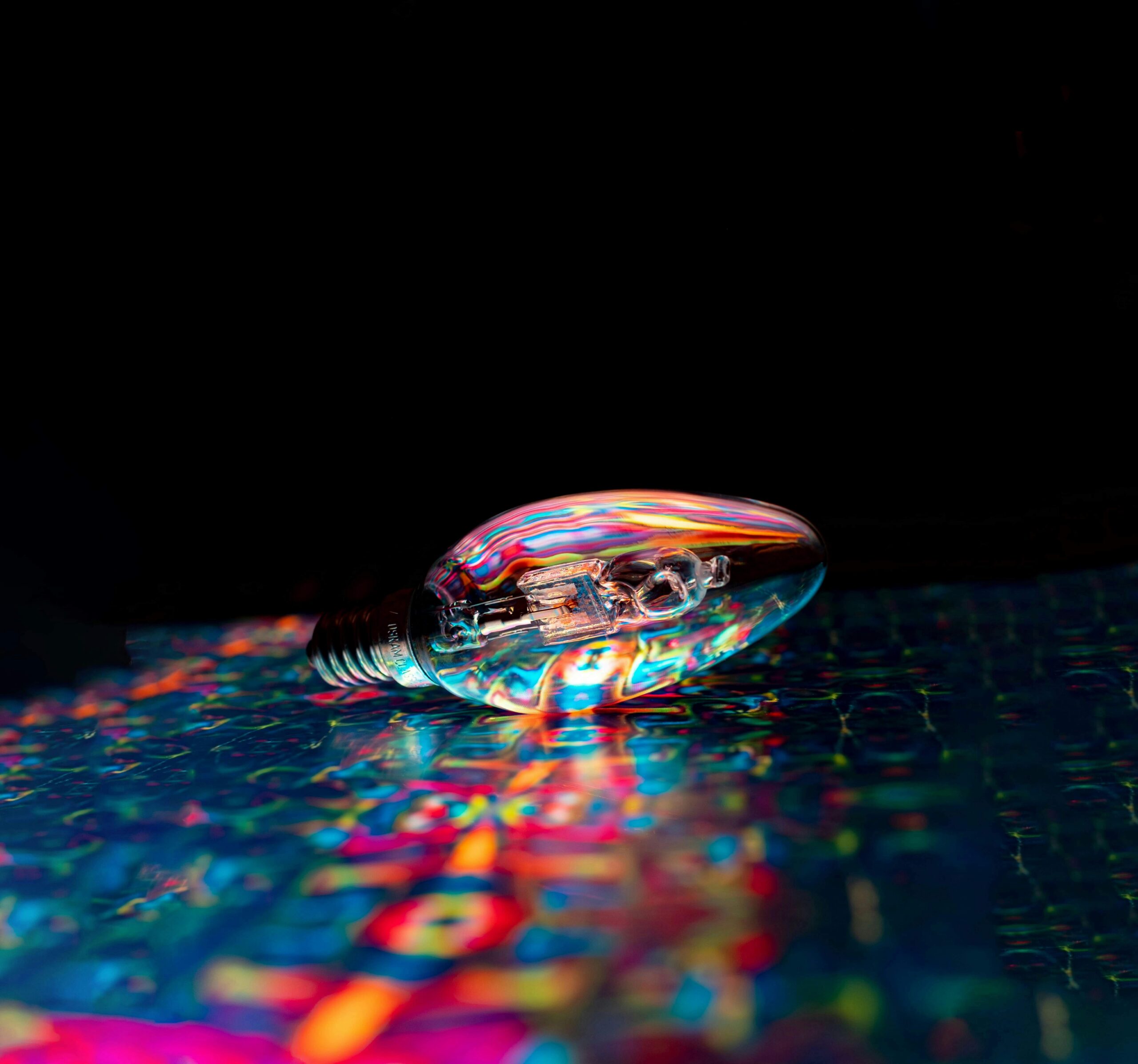 Close-up of a reflective light bulb lying on a colorful illuminated surface against a dark background, with vibrant rainbow-like reflections symbolizing creativity and innovation.